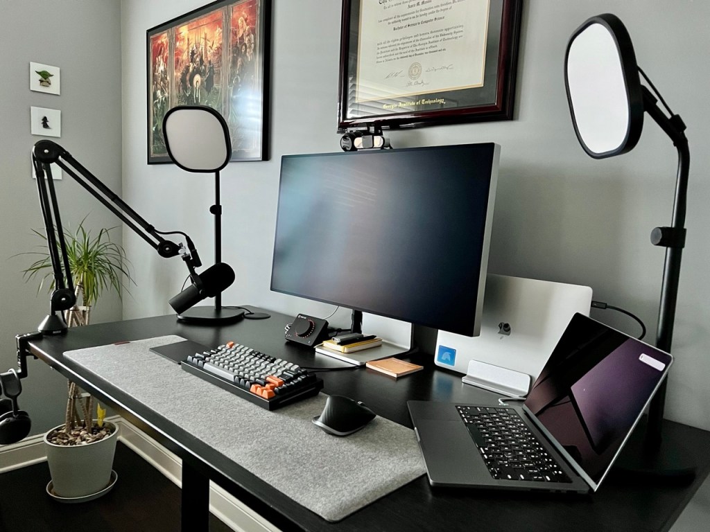 A mostly wire free desk aimed from the right side showing various work equipment: a large monitor, a small laptop to the right and one behind in a stand, two key lights hovering over the desk, a microphone, webcam, keyboard, mouse, trackpad, volume knob, and a notebook, pen, and more